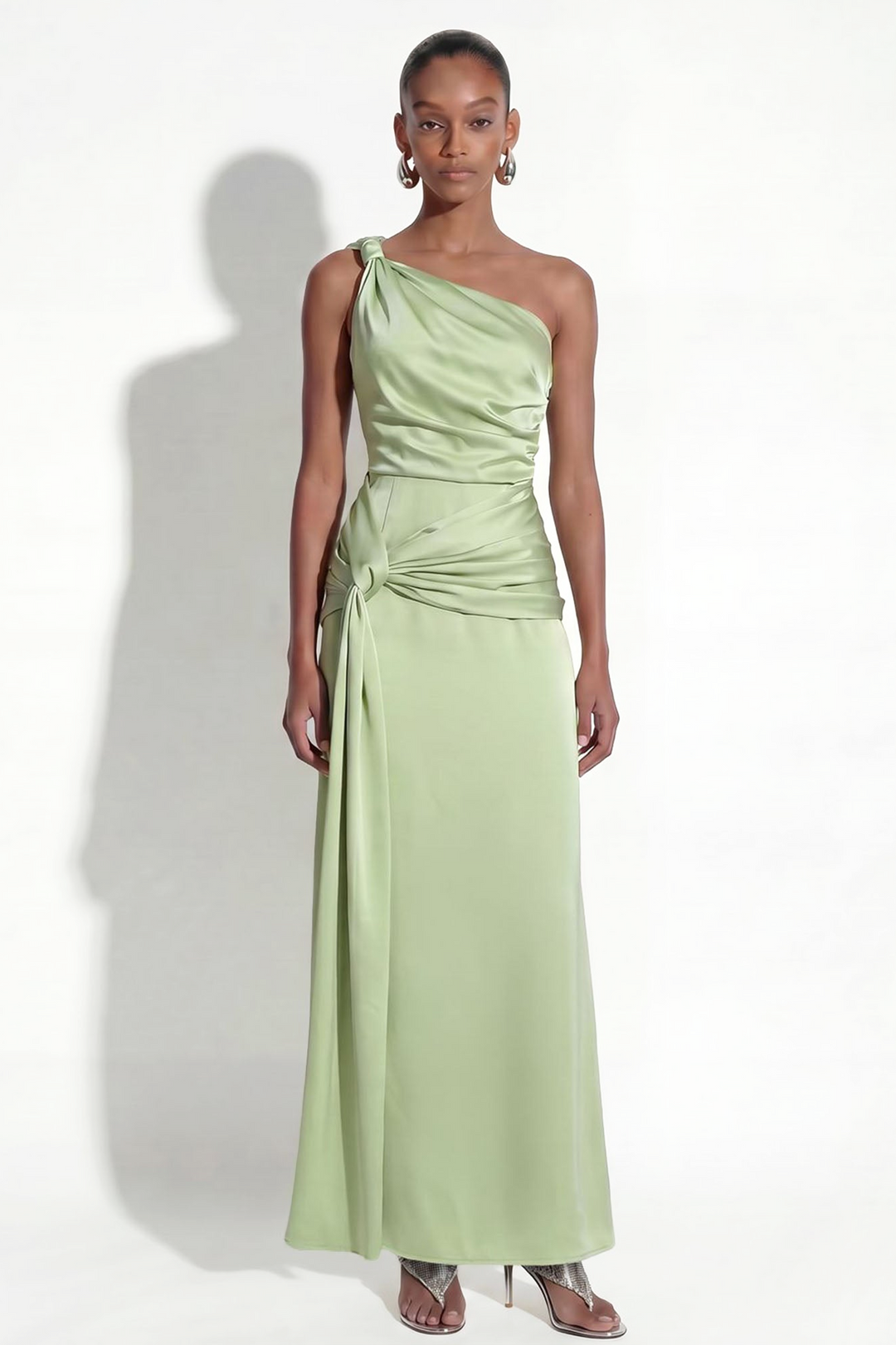 A model wears a sage green one-shoulder satin maxi dress with a gathered, twisted waist detail, paired with metallic silver high-heeled sandals against a white studio background.